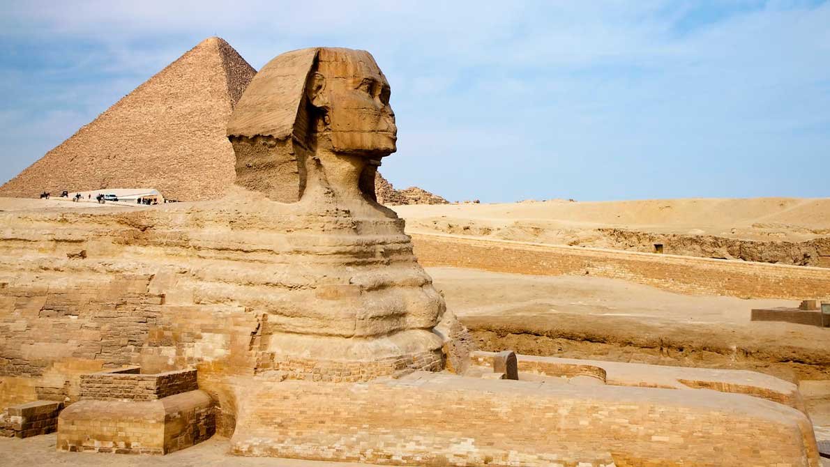 Передний боковой ракурс Сфинкса с пирамидой Хеопса, видимой на заднем плане пустынного плато.